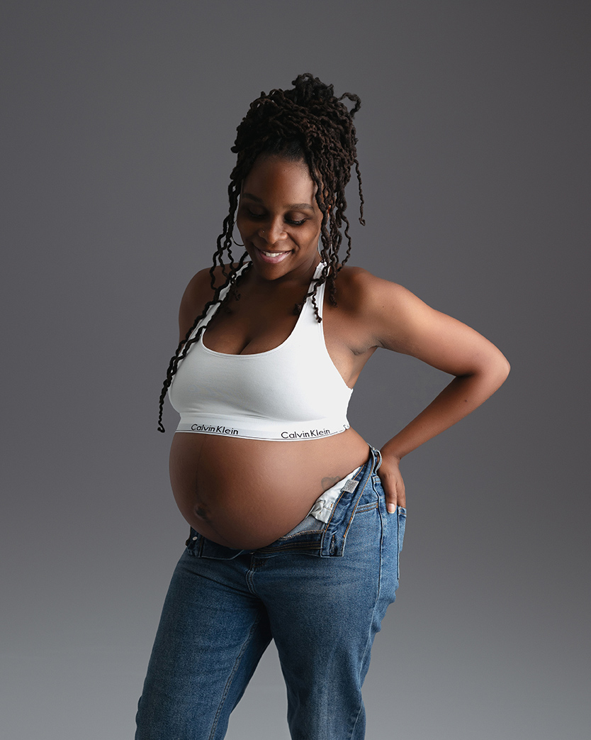 pregnant women poses with hand on hip while she smiles at her belly in white bralette and jeans