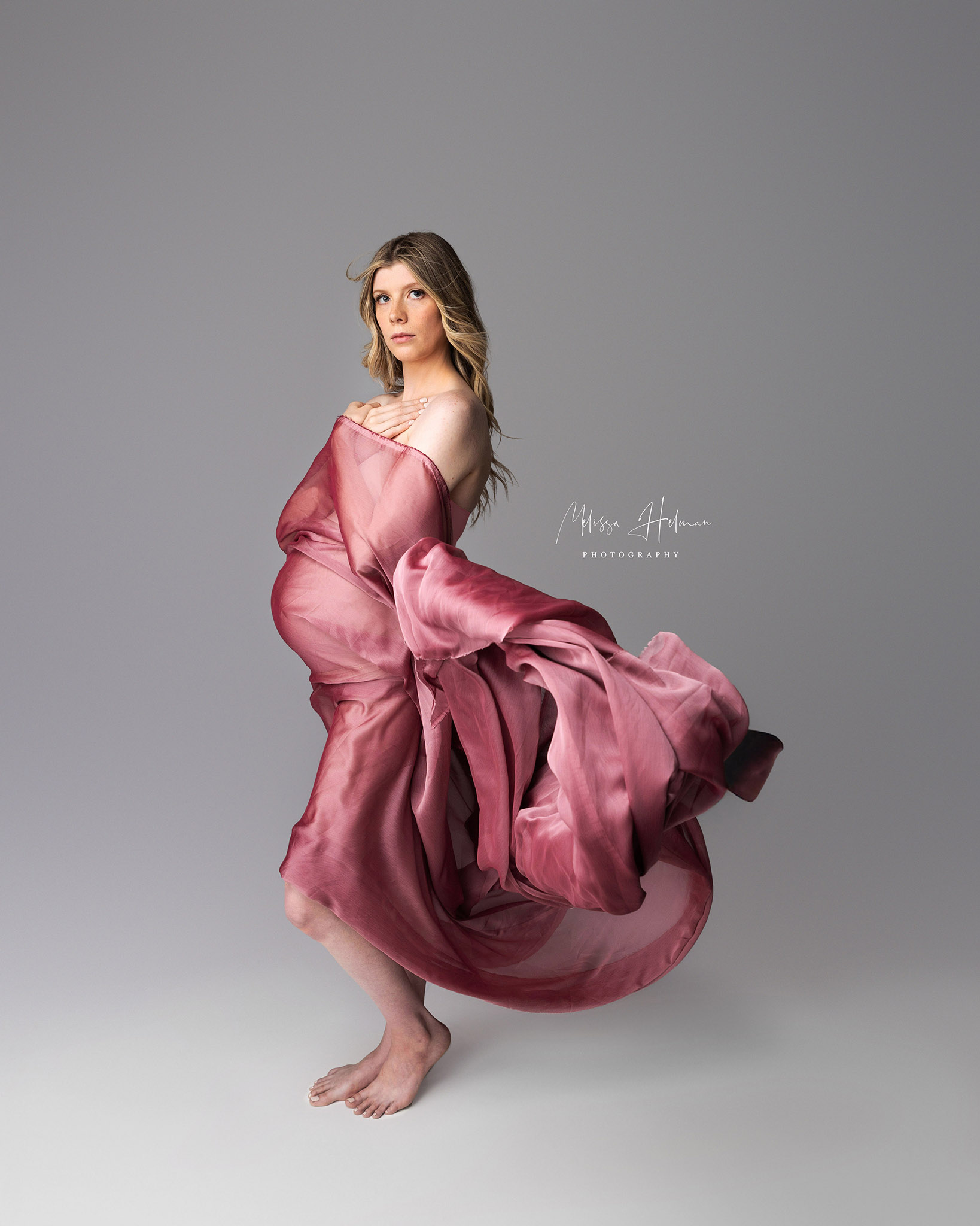 pregnant woman sting with pink fabric and wind blowing.