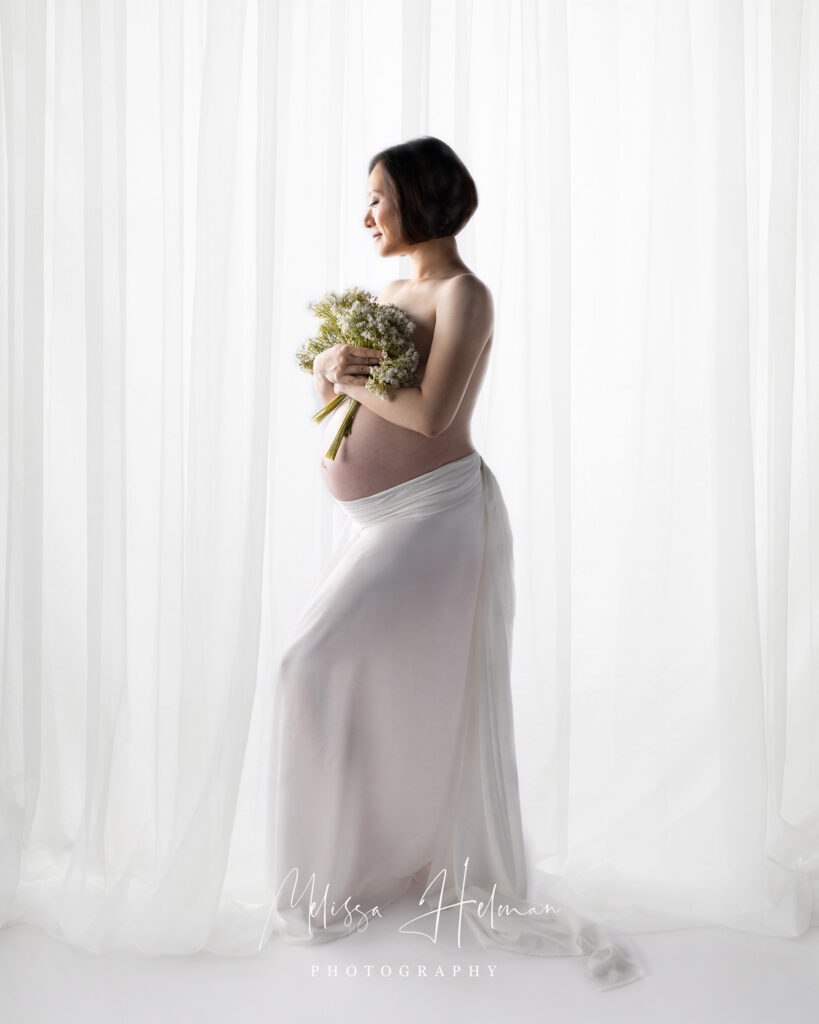 Mother posing with flowers and silk white skirt for maternity portrait with sheer curtain behind.