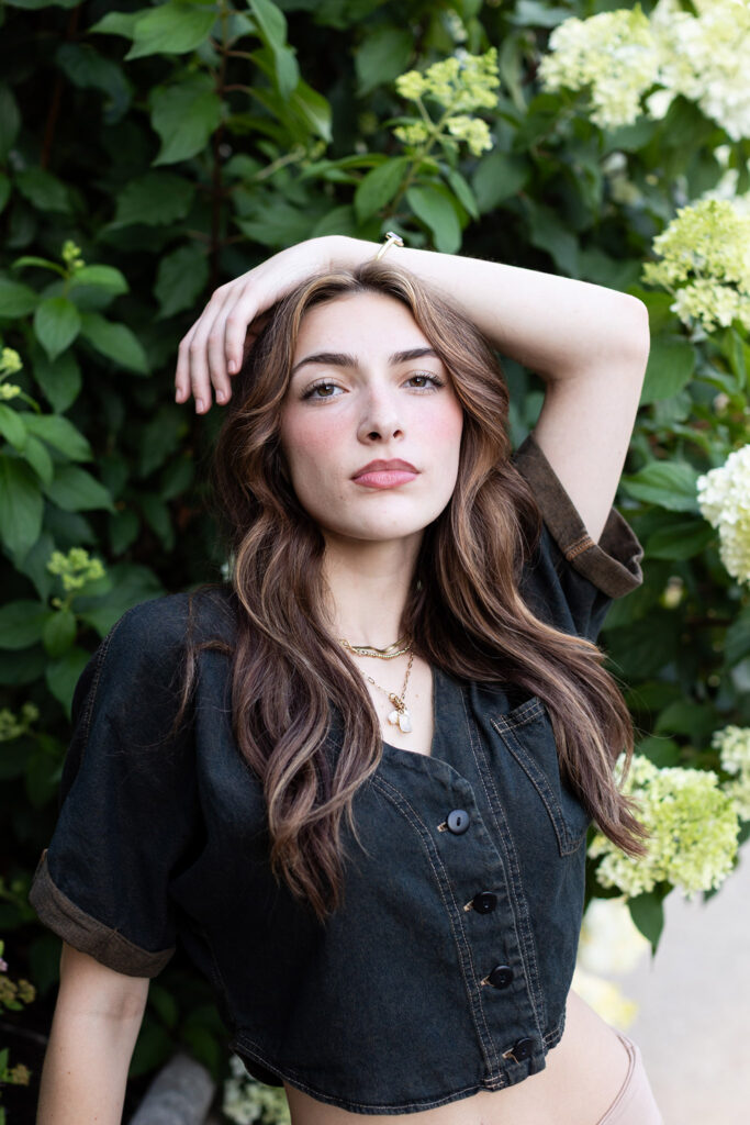 Girl poses with hand on head near white flower bush for senior portraits.